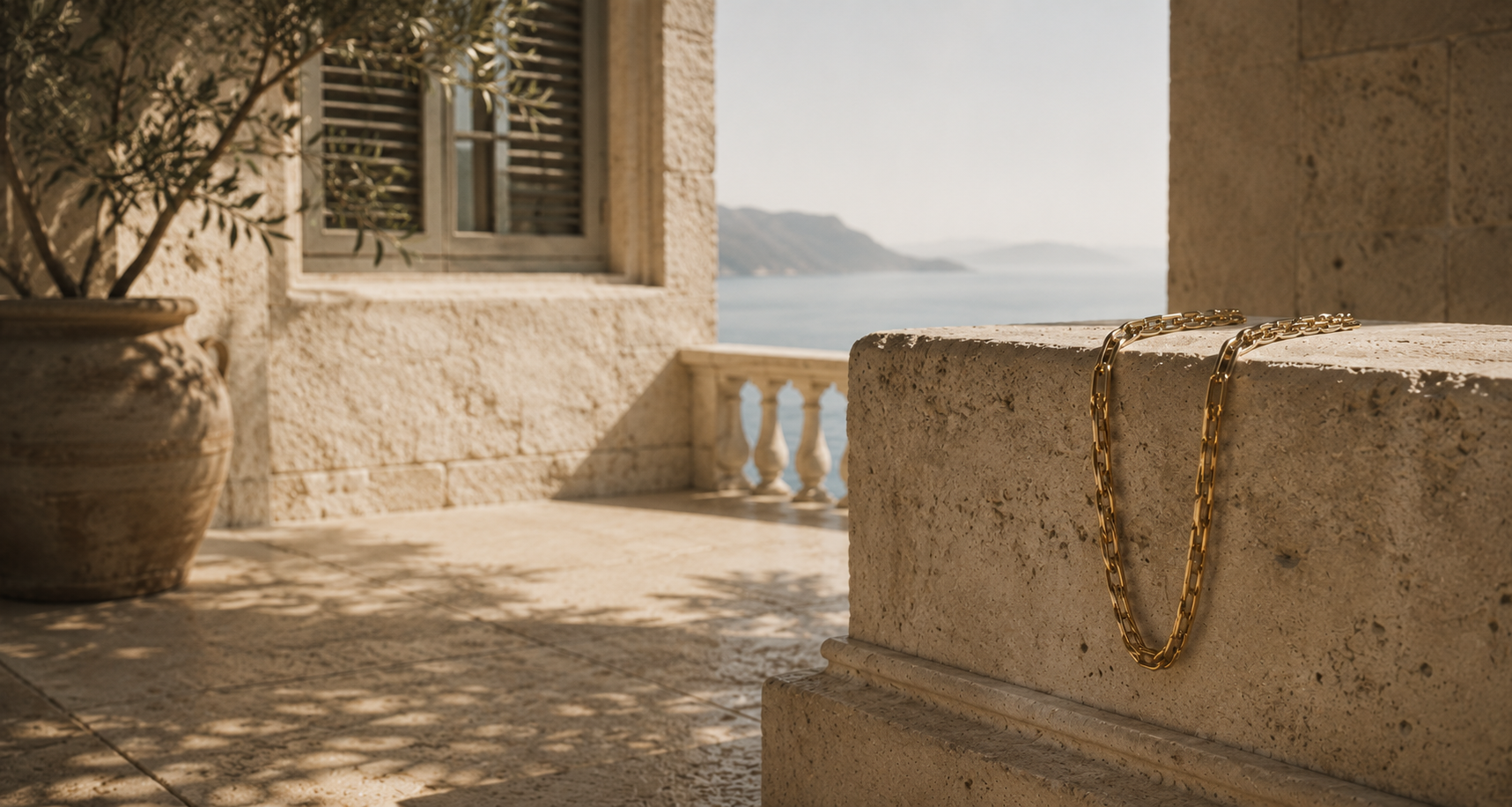 A gold chain necklace draped over a beige stone wall in an outdoor Mediterranean setting with a sea view in the background.